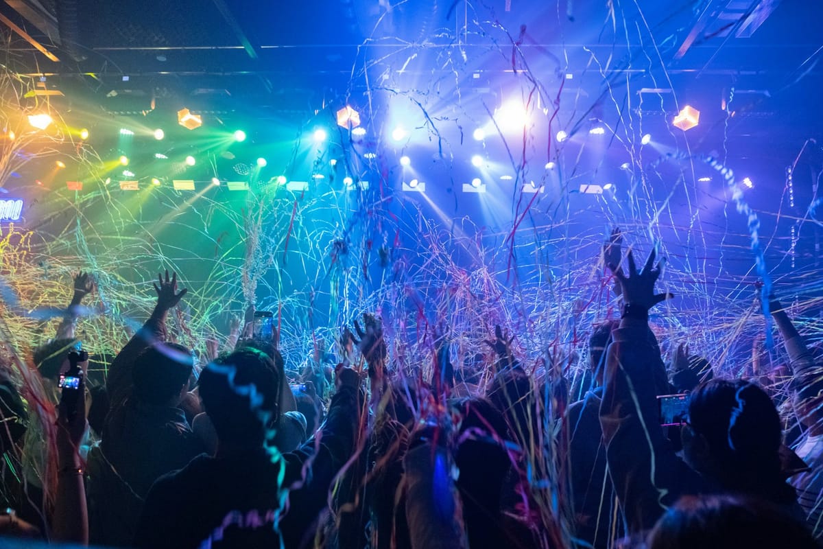 A crowd with hands raised under colorful stage lights and streaming confetti